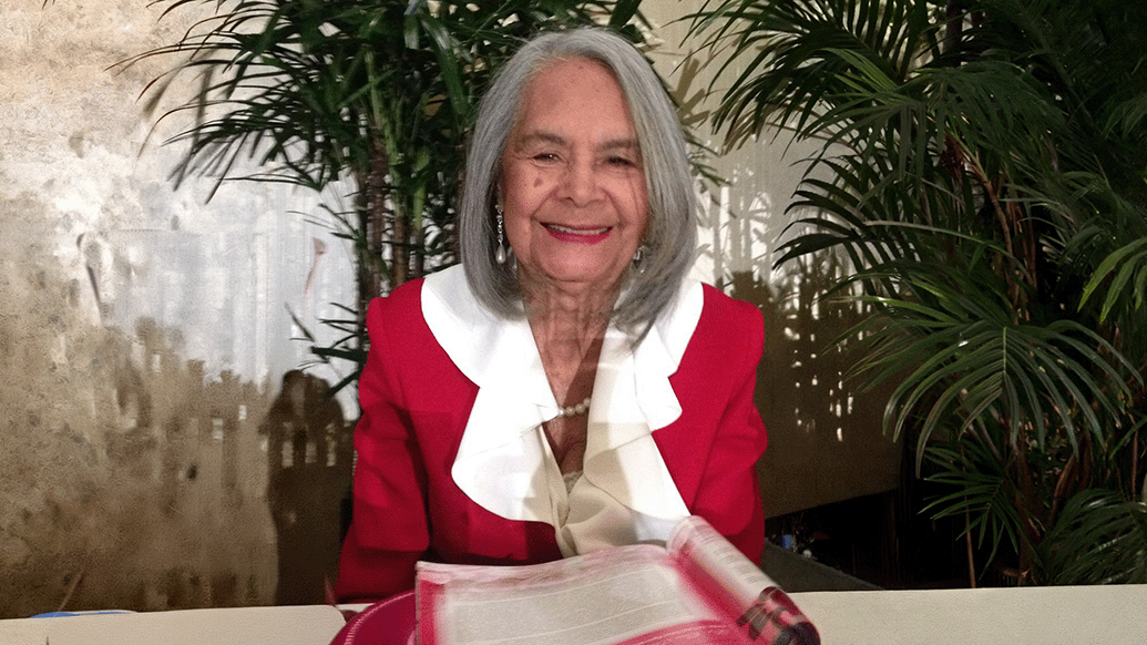 Photograph of an elderly woman wearing a red jacket with a large white ruffled collar, sitting at a table with an open book in front of her. Background includes green leafy plants and a textured wall, suggesting an indoor setting with natural light.