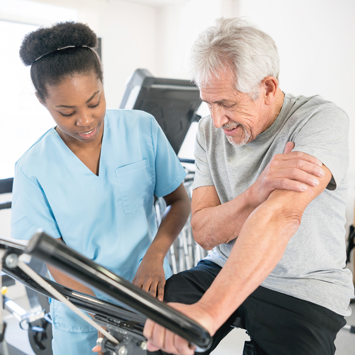 Healthcare worker in scrubs assists an elderly person using an exercise machine in a clinical setting.